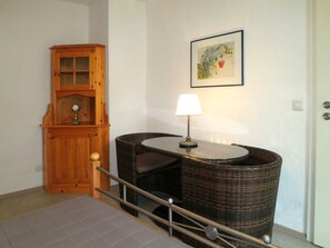 Una stanza con una sedia in vimini, un tavolino con una lampada, un armadietto in legno e un quadro appeso al muro.