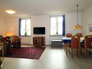 Una sala de estar con televisión, una mesa de comedor con sillas, un tapete rojo estampado y una ventana con cortinas.