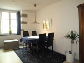 Ein Essbereich mit Tisch, Stühlen, einer blauen Tischdecke, einem gemusterten Teppich, einer Topfpflanze und einem Fenster mit Vorhängen.