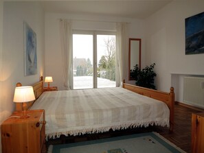 Un dormitorio con una cama grande, dos lámparas de noche, una ventana con cortinas, un espejo, un radiador y un cuadro en la pared.