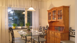A dining area with a glass table, metal chairs, a wooden cabinet with glass doors, and a window with curtains.