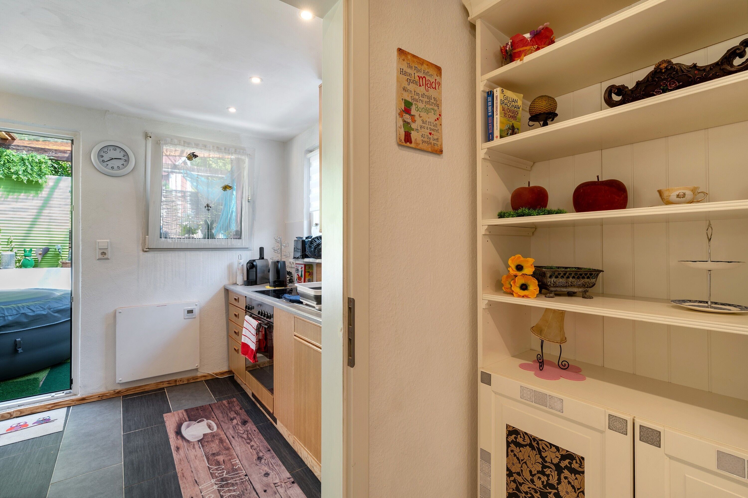 A compact kitchen with a clock, a radiator, and a window with a view of greenery.