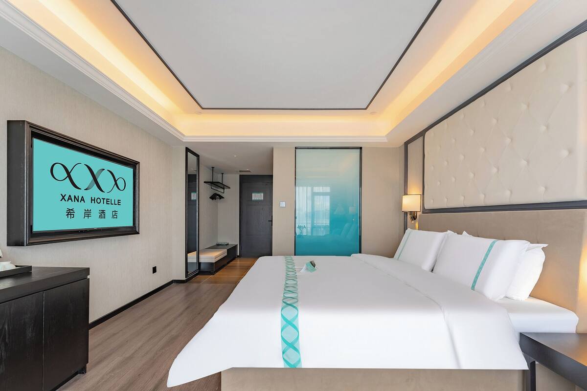 A modern hotel room with a large bed, a bedside table, a desk, and a TV displaying the hotel name.