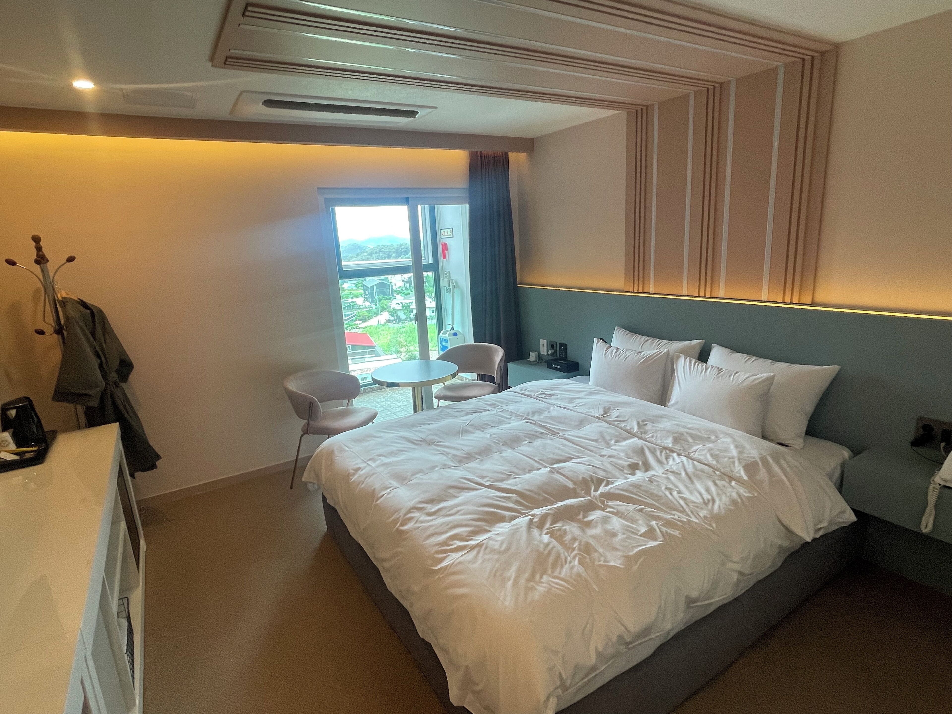 Quarto de hotel com uma cama grande, uma mesa pequena, duas cadeiras, uma escrivaninha com telefone e vista para o exterior através de uma janela.