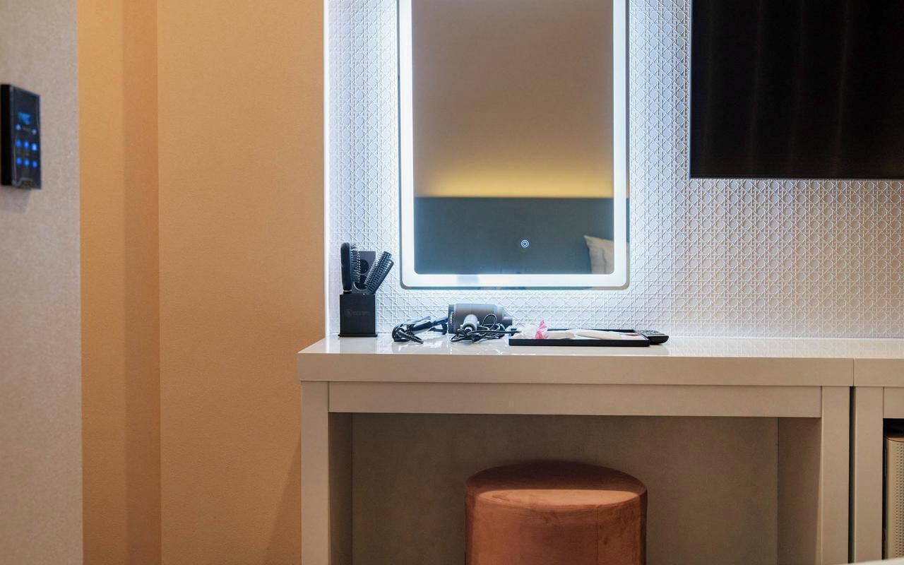 Una habitación de hotel moderna con un escritorio, un taburete pequeño, una ventana con vistas y un panel de control en la pared.