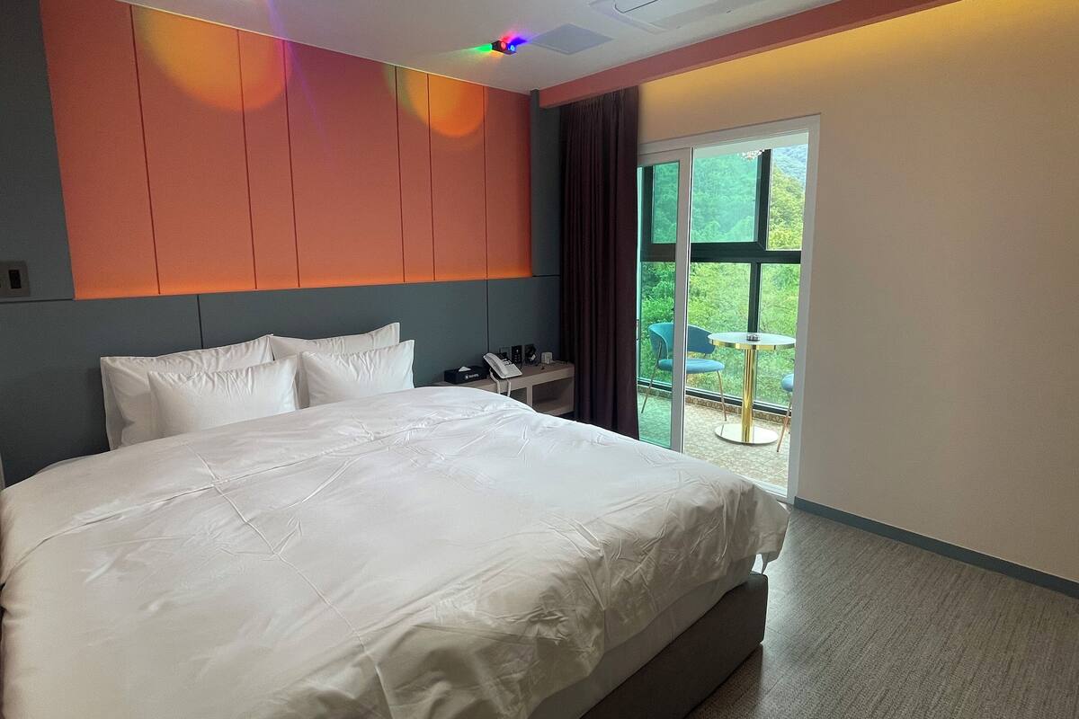 A hotel room with a large bed, a bedside table with a phone and a box, and a view of greenery through the window.