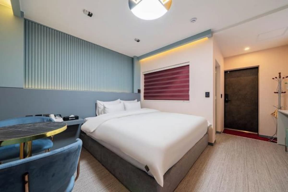 A hotel room with a bed, a desk, and a chair. There is a window with blinds, a door, and a coat rack.