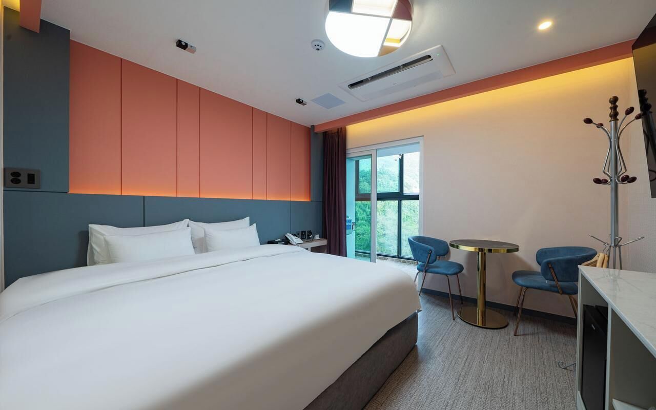 A modern hotel room with a large bed, a desk with a chair, and a window with a view of greenery.