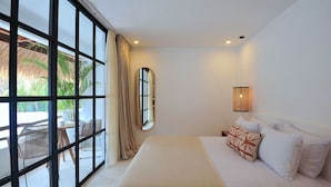 A hotel room with a large window, a bed with white linens, a hanging light fixture, and a view of a thatched-roof structure outside.