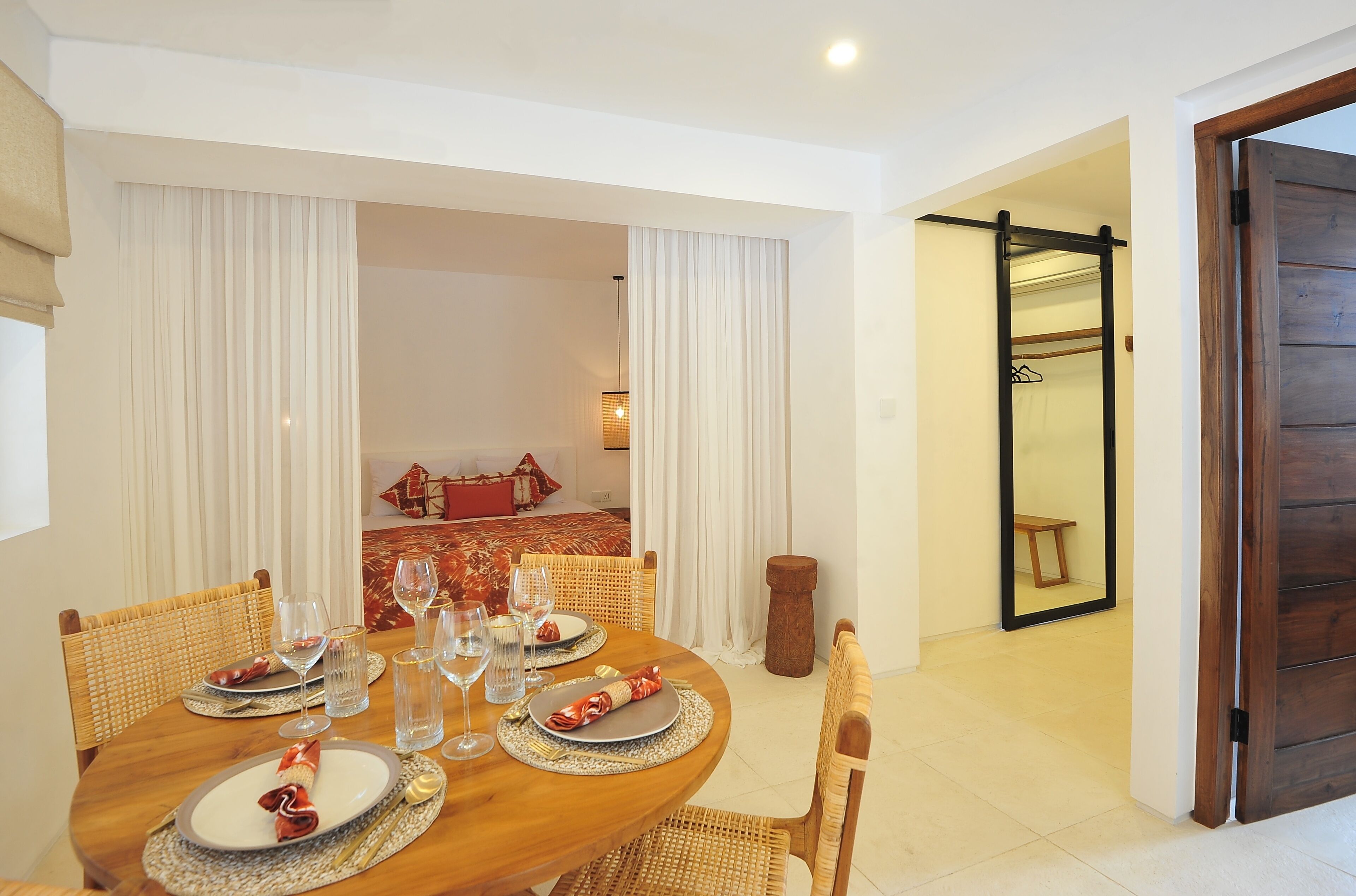 A dining area with a wooden table set for four, surrounded by wicker chairs. A small wooden stool is placed near the table.