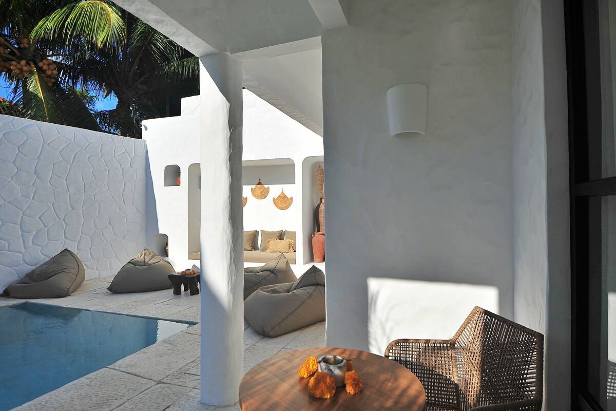 A modern outdoor lounge area with a pool, seating, and a view of a white building with a textured wall and hanging lights.