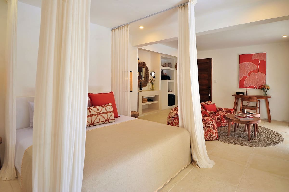 A bedroom with a large bed, a desk, and a chair. There is a wardrobe and a shelf with decorative items.