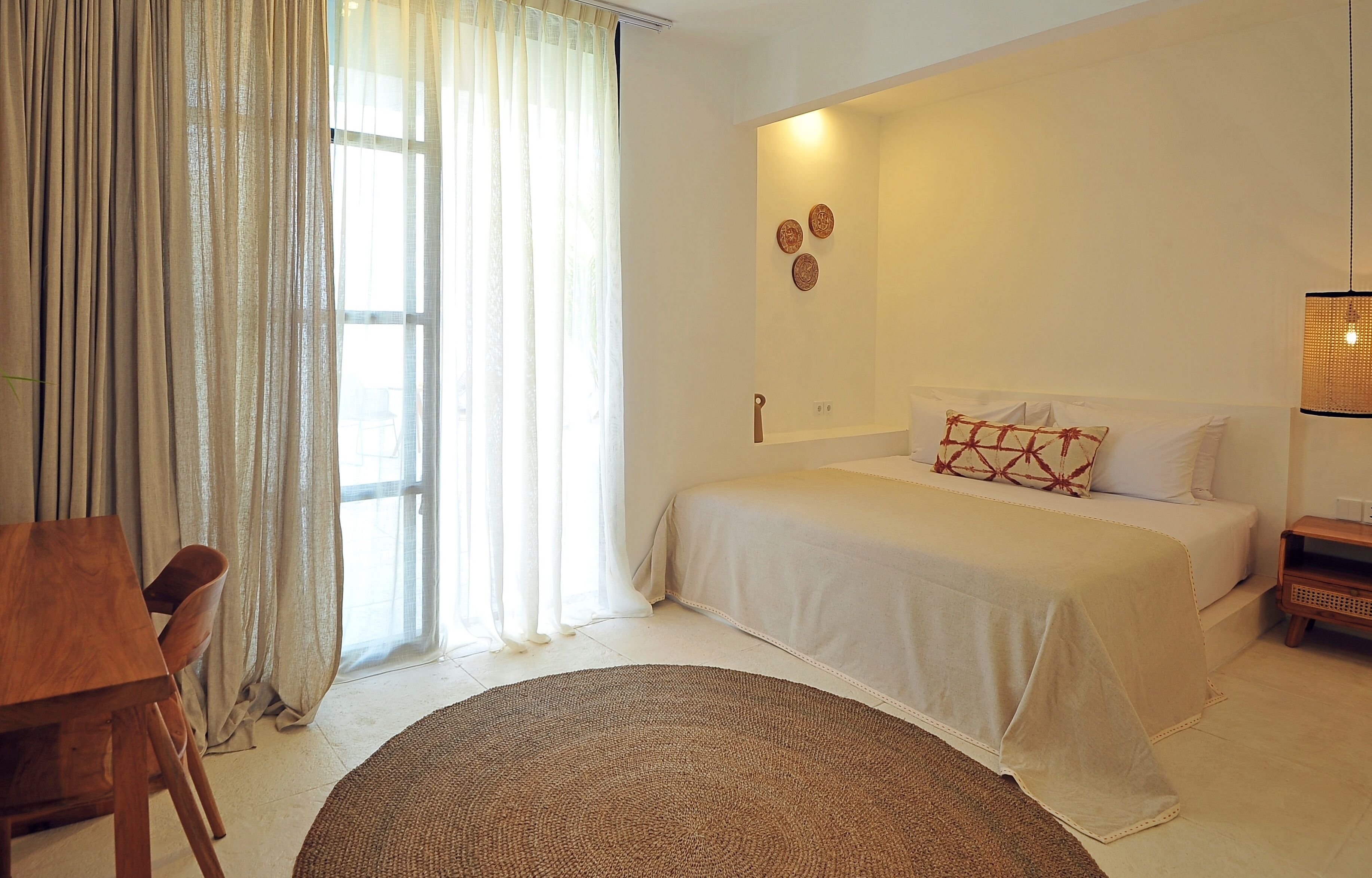 Un dormitorio con cama, una mesita de noche, una silla, un ventanal con cortinas y una lámpara de pared.
