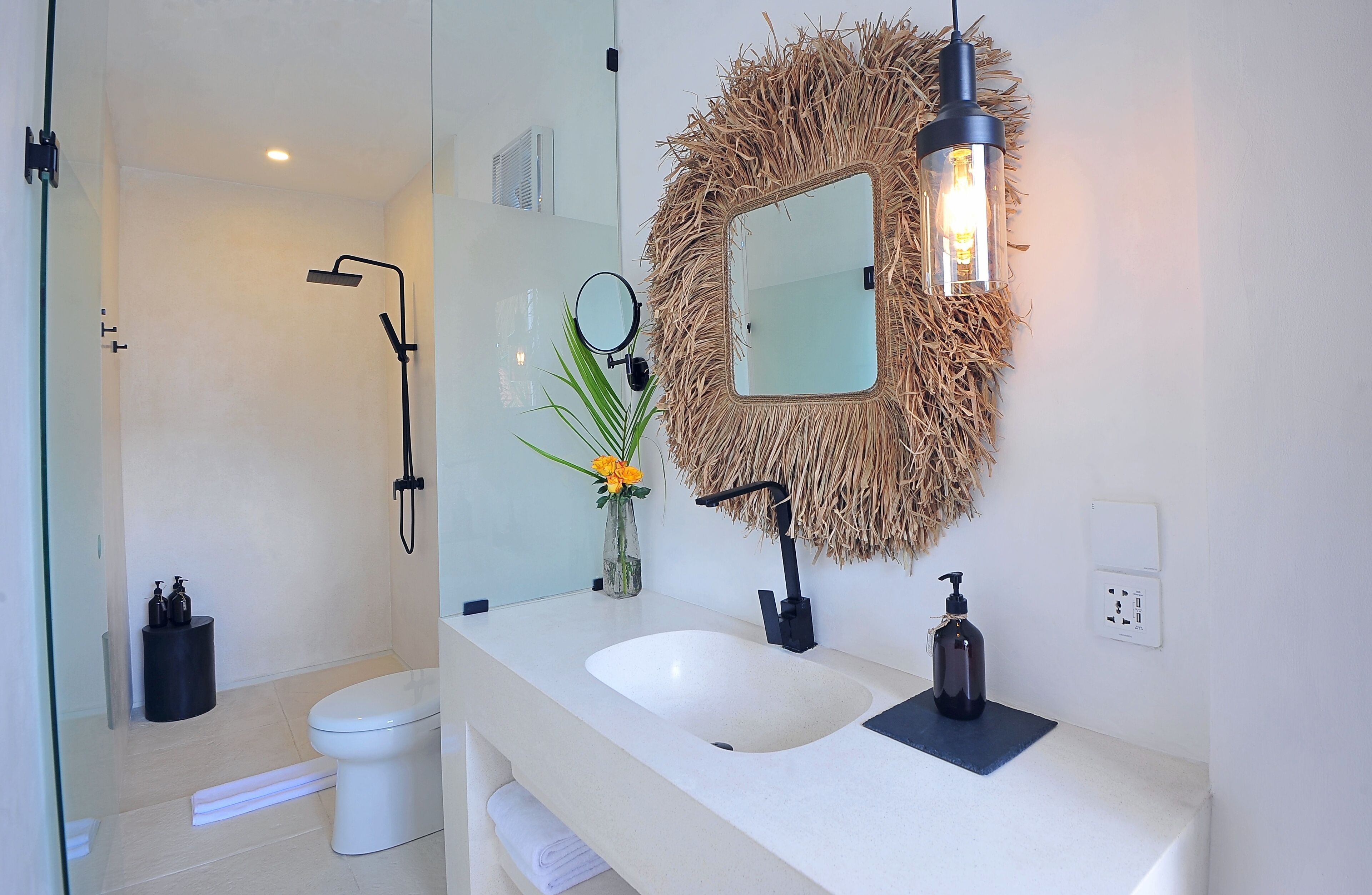 A modern bathroom with a white sink, a round mirror with a straw-like frame, and a handheld showerhead.