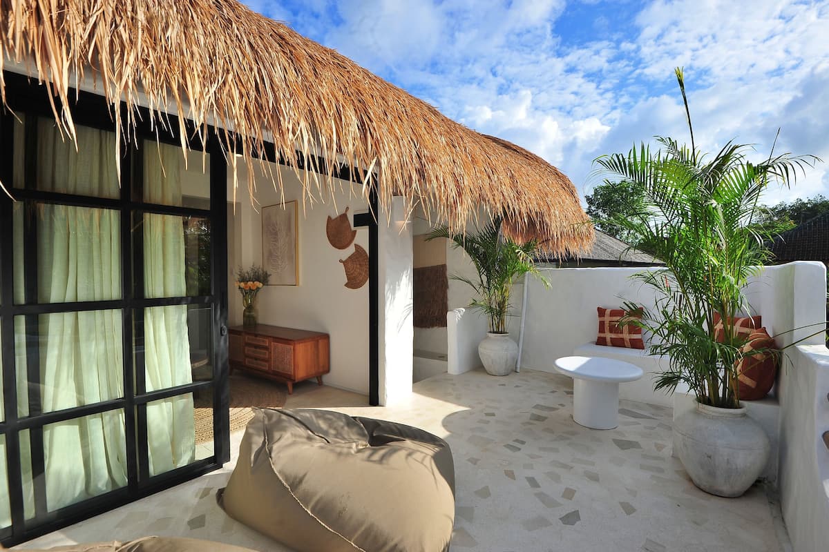 A modern outdoor living area with a thatched roof, a large sliding glass door, a cushioned seating area, and potted plants.