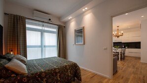 A bedroom with a bed, a window with curtains, a picture on the wall, and a kitchen in the adjacent room.