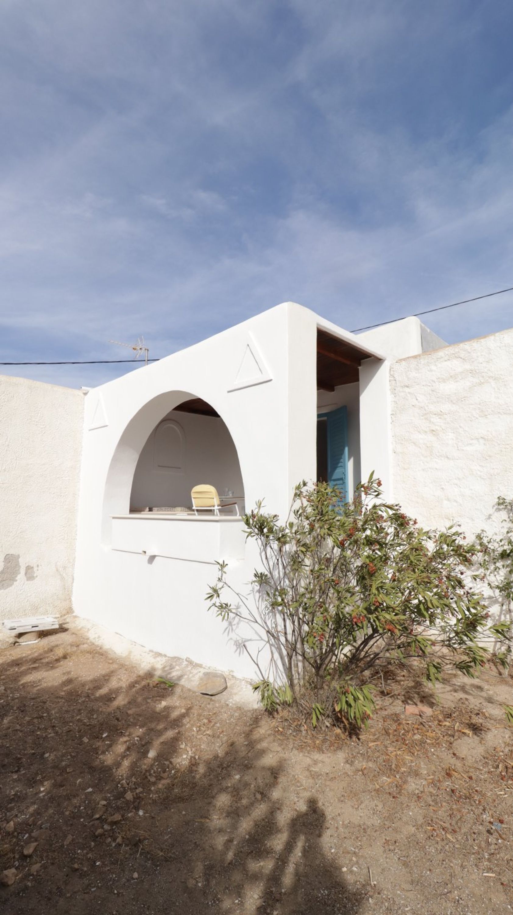 Une petite structure blanche en forme d’arche, avec un banc à l’intérieur, une porte et une petite plante devant.
