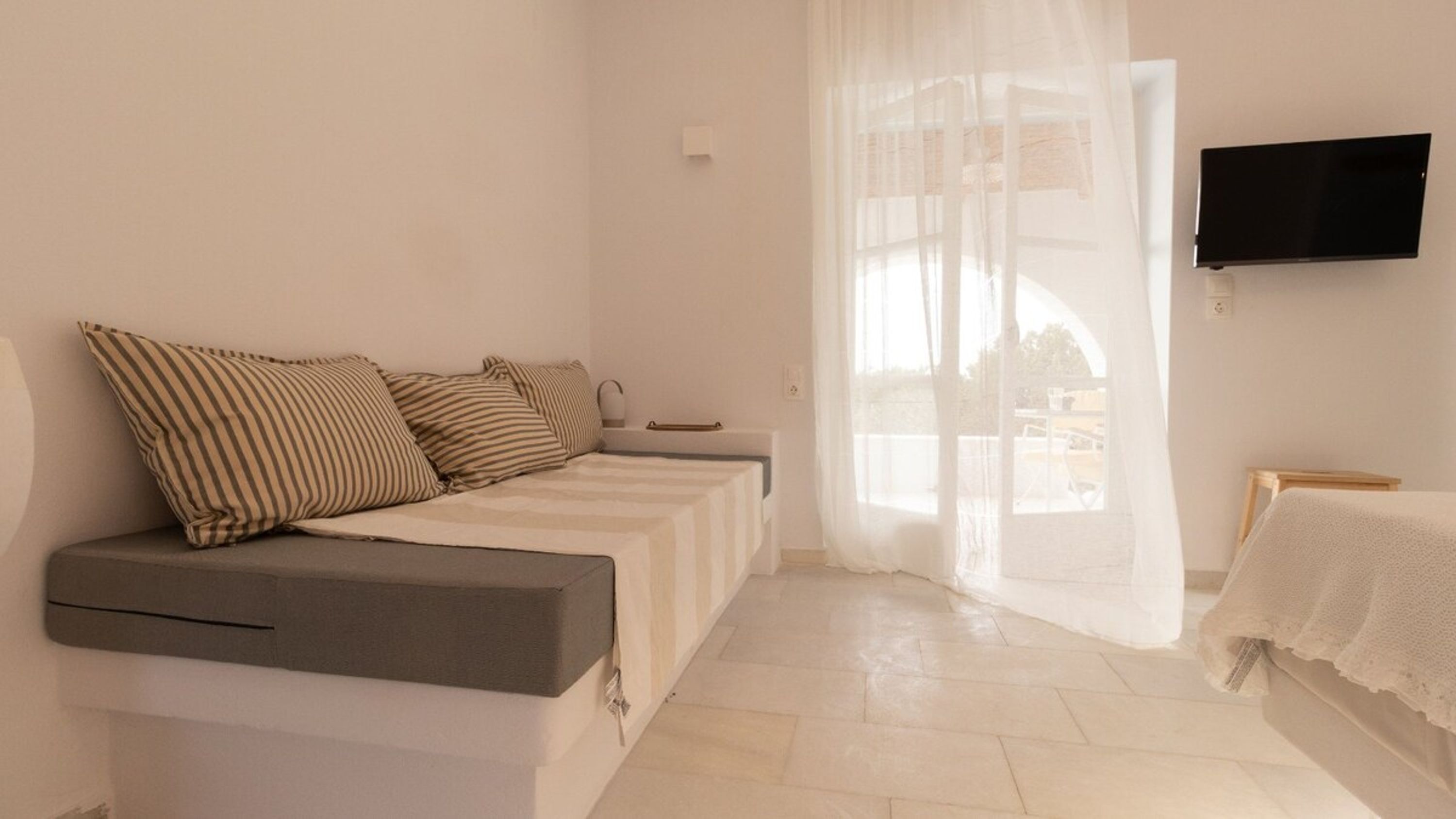 Une chambre moderne dotée d’un canapé-lit, d’un téléviseur à écran plat fixé au mur et d’une grande fenêtre agrémentée de rideaux légers.