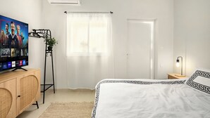 A bedroom with a bed, a TV on a wooden cabinet, a potted plant, a window with curtains, and a bedside table with a lamp. - Florentin Stylish Stay by Sea N' Rent (Tel Aviv)