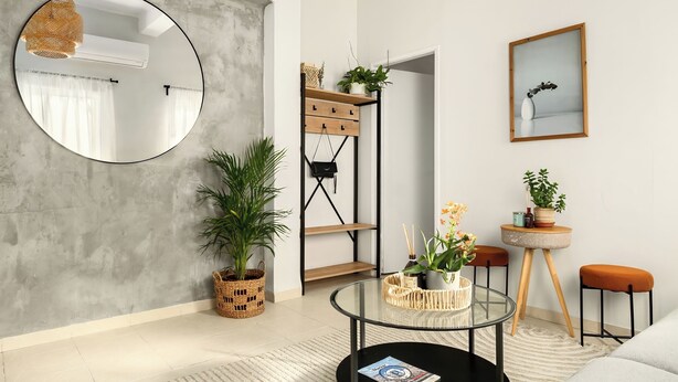 A modern living room with a round mirror, a glass coffee table, a wicker side table, and a wooden shelf with plants. - Florentin Stylish Stay by Sea N' Rent (Tel Aviv)