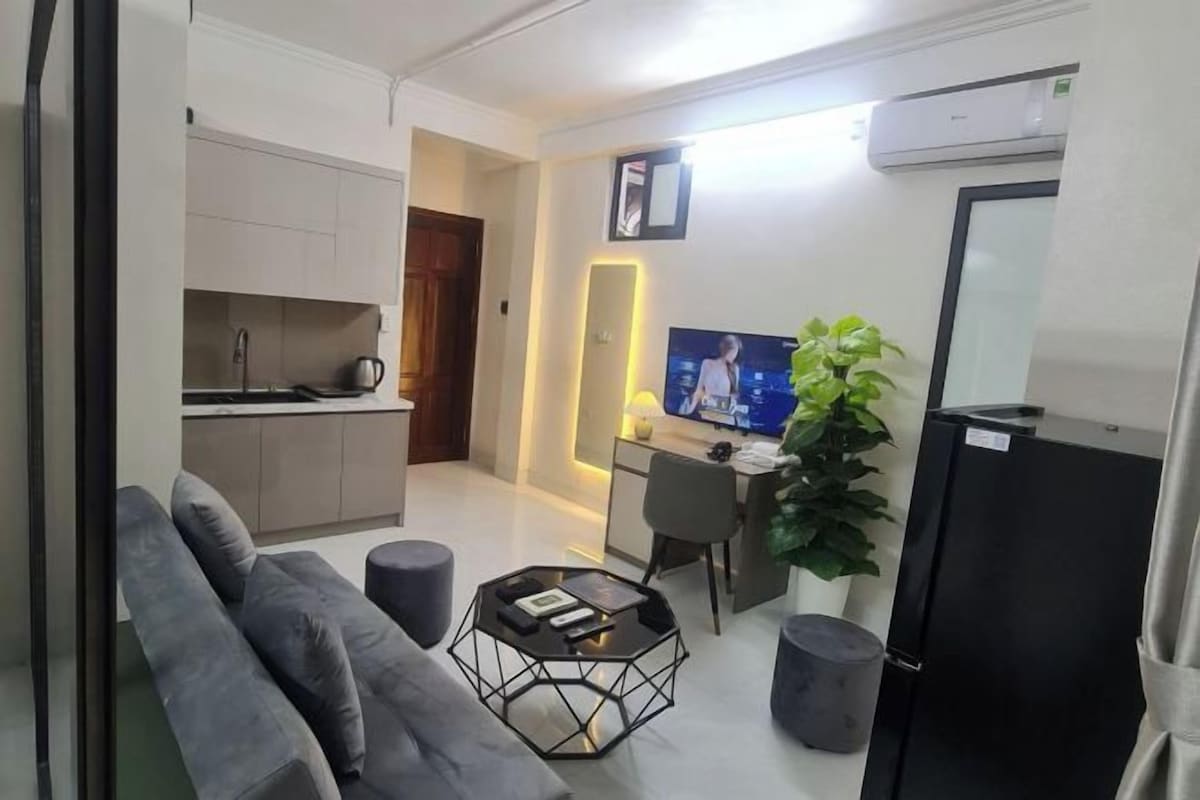 A modern living room with a sofa, coffee table, and a TV. There's a kitchen area with cabinets and a refrigerator.