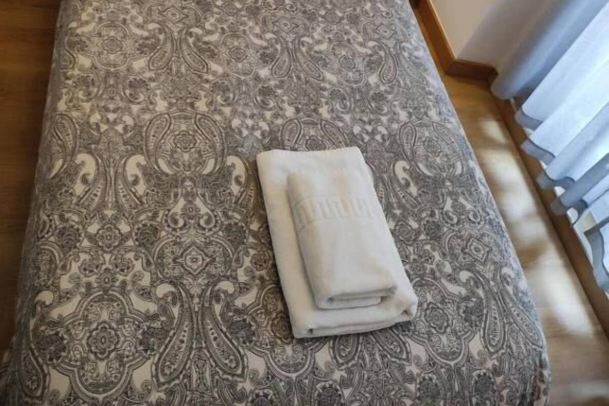 Una cama bien hecha con una colcha estampada y una toalla doblada, una mesita de noche con un tel éfono, y una pared con un interruptor de luz.