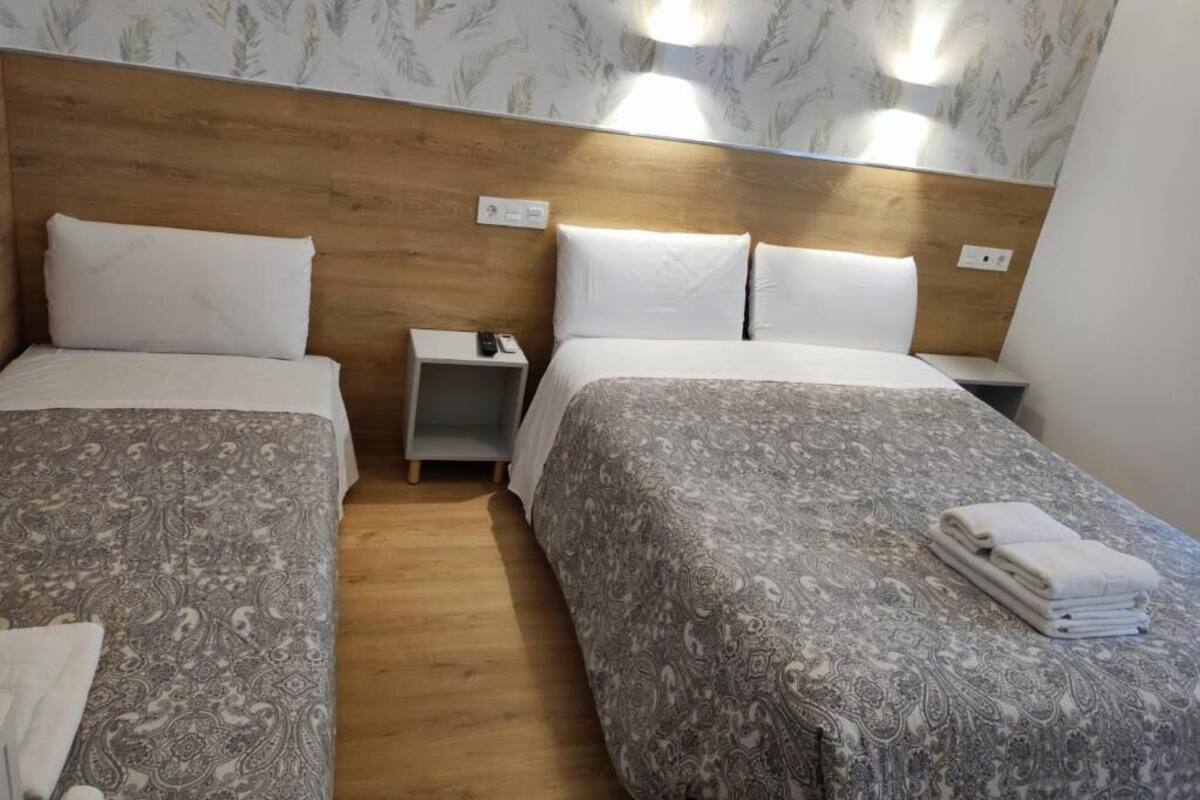 Habitación de hotel con dos camas, cabecero de madera, ropa de cama estampada, mesitas de noche e iluminación montada en la pared.