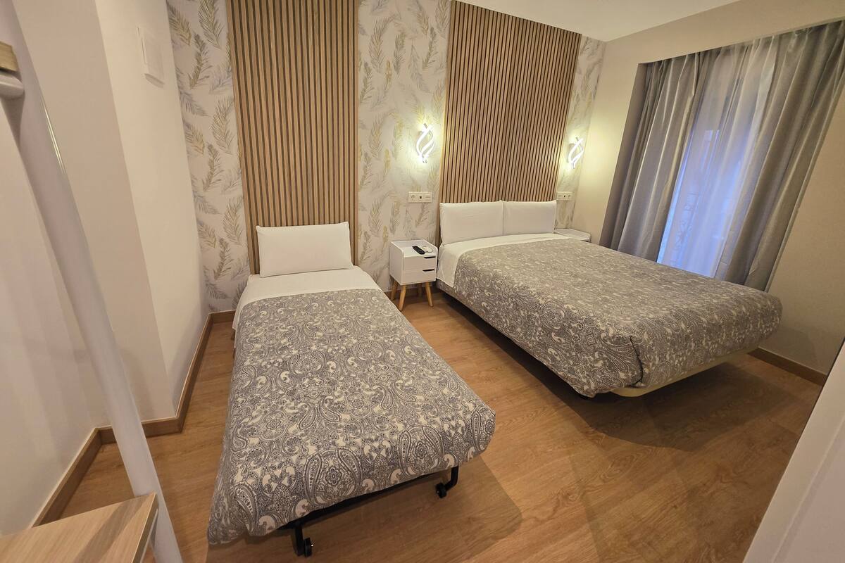 Habitación de hotel con dos camas, cabecera de madera, mesita de noche e iluminación moderna.