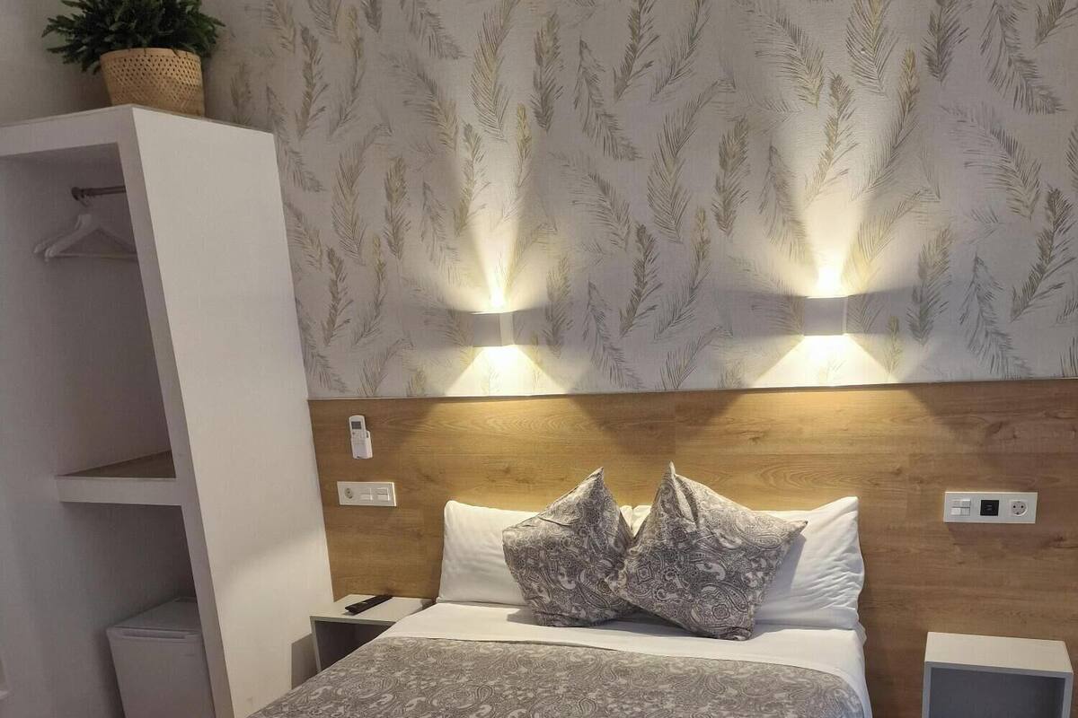 Una cama bien tendida con ropa de cama estampada, una mesita de noche con una lámpara y un estante en la pared con una planta.