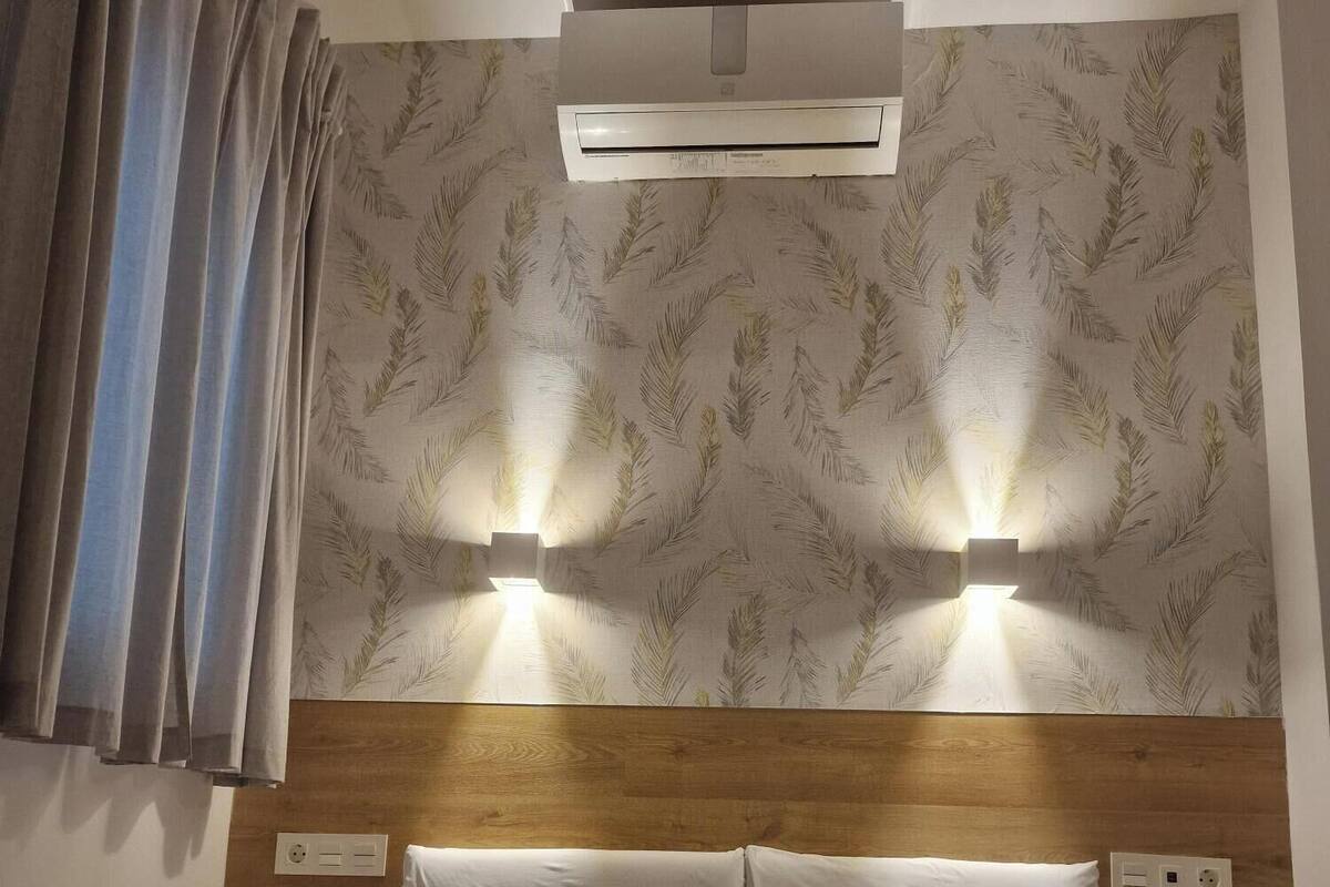 Habitación de hotel con cabecera de madera, dos luces fijadas a la pared, colcha estampada y una pila de toallas dobladas.