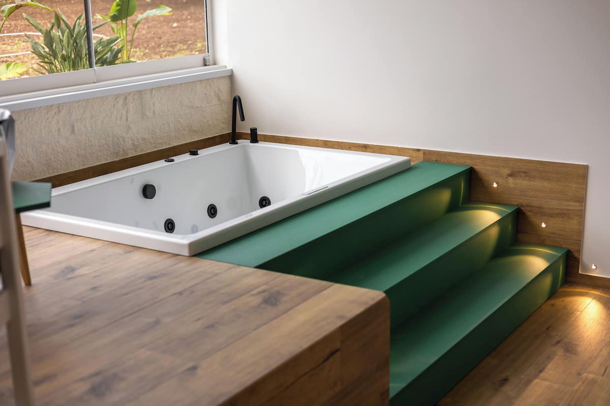 A modern bathroom with a freestanding bathtub, wooden steps, and a window with a view of greenery.