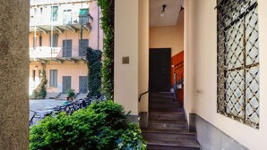 Una escalera estrecha que lleva a una puerta, con una ventana de celosía metálica a la derecha y un edificio con balcones al fondo.