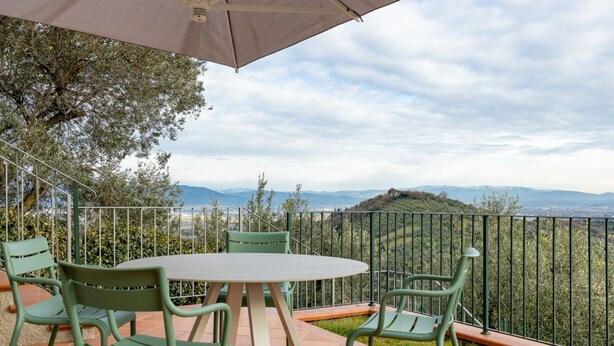 Une terrasse avec une table ronde et des chaises, une rambarde en métal et une vue sur des collines lointaines.