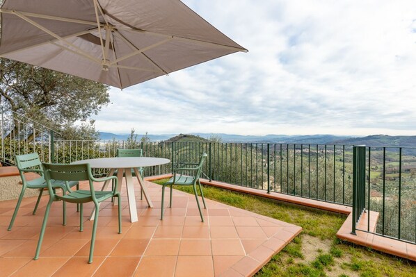 Une terrasse avec une table et des chaises, un grand parasol et une vue sur des collines lointaines.