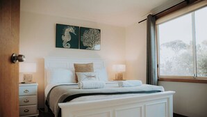 A bedroom with a white bed, two bedside tables with lamps, a window with curtains, and two wall art pieces. - Spindrift Falmouth, adventurers coastal haven (Break O'Day)