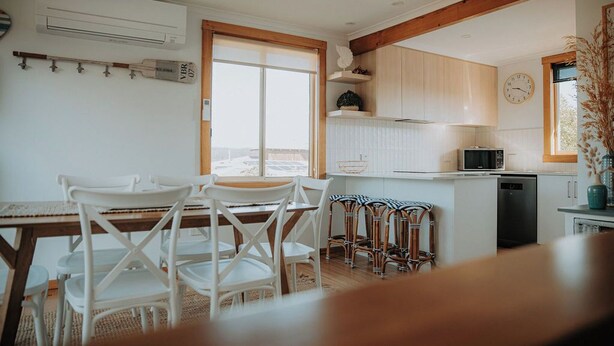 A modern kitchen with white cabinets, a dining area with white chairs, and a large window. - Spindrift Falmouth, adventurers coastal haven (Break O'Day)