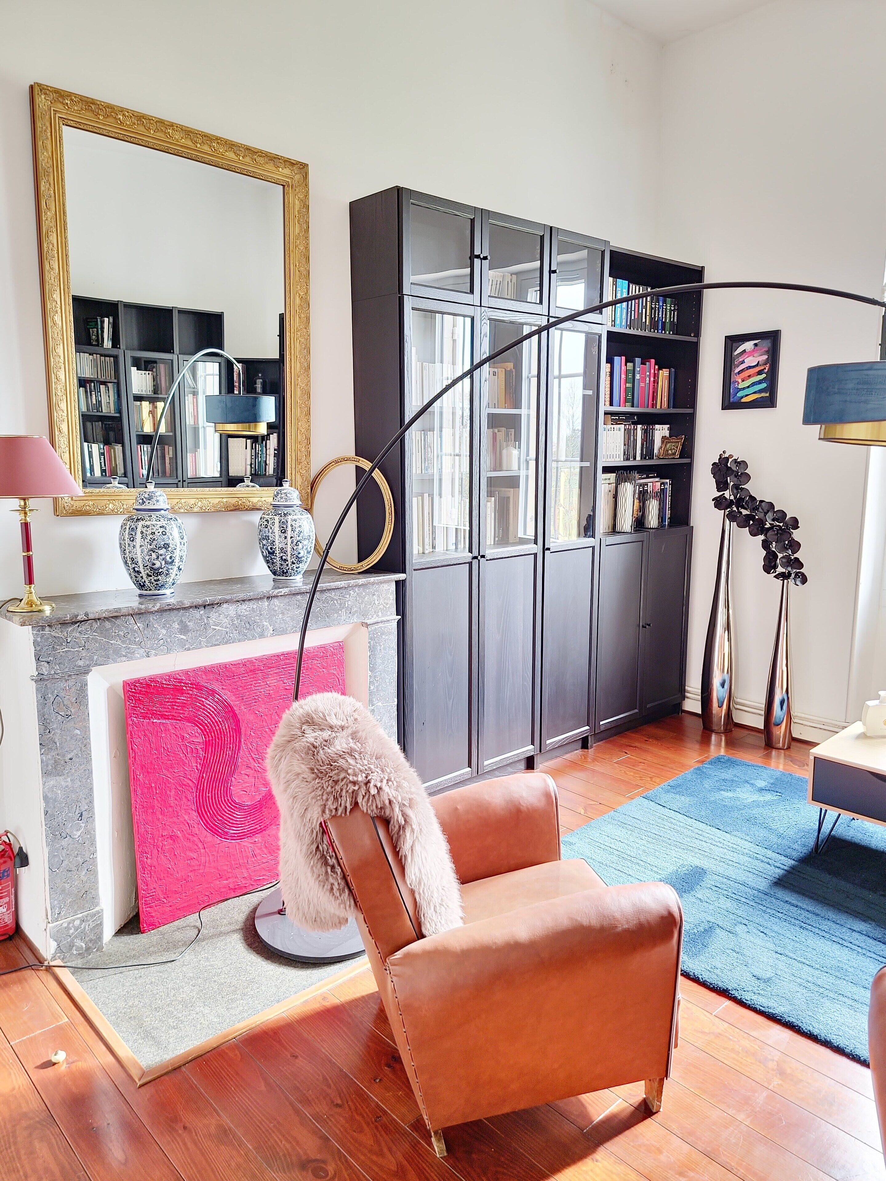 A modern living room with a fireplace, a leather armchair, a bookshelf, and a large mirror.