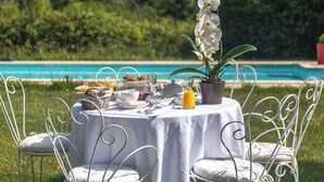 A dining table set outdoors with white chairs, a tablecloth, and a view of a swimming pool in the background. - MANOIR LE COUTUREAU (Rouffignac-de-Sigoulès)