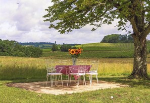 A table set outdoors with a red and white checkered tablecloth, surrounded by white chairs, under a tree with a vase of sunflowers. - MANOIR LE COUTUREAU (Rouffignac-de-Sigoulès)
