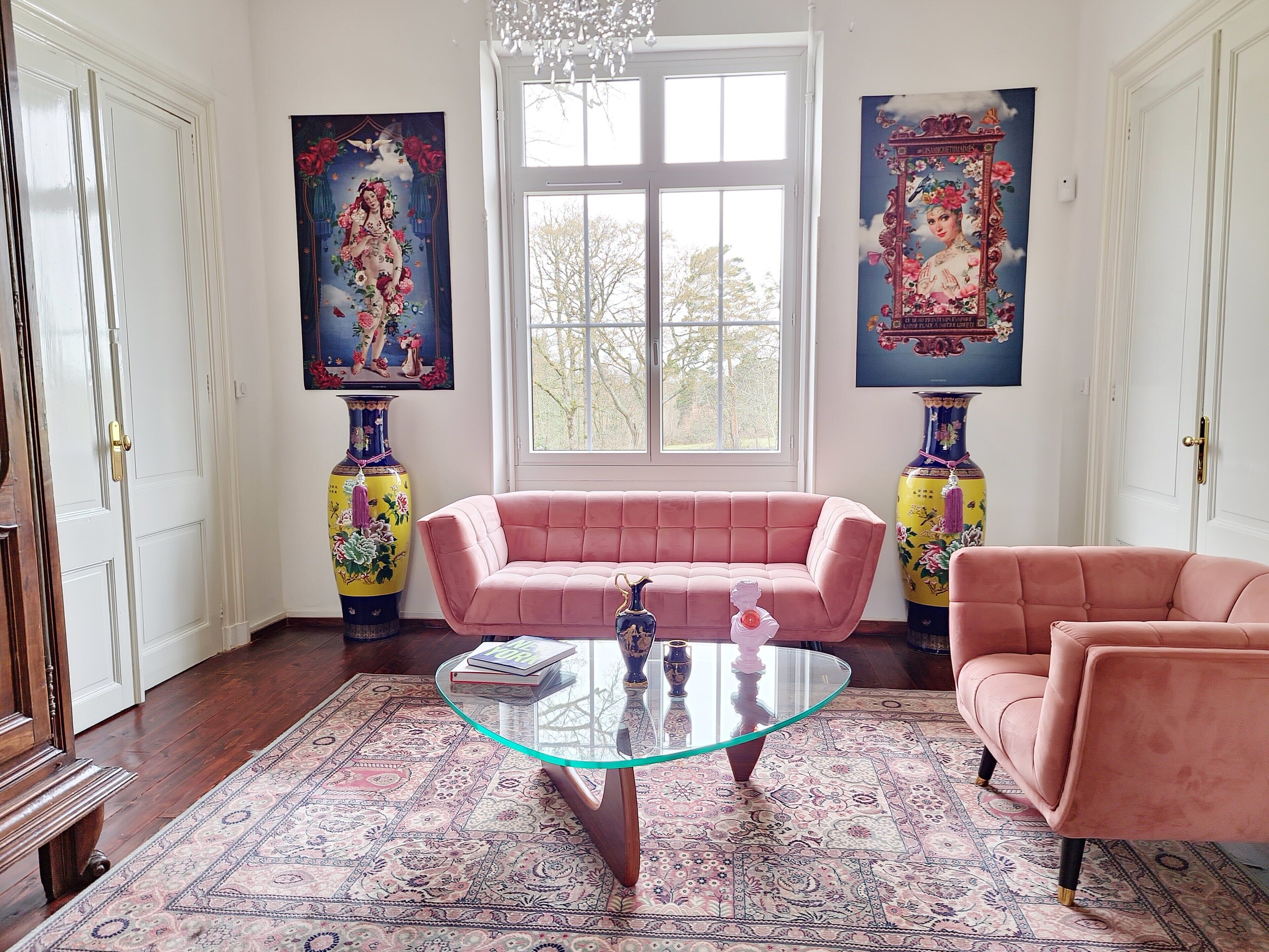A room with a pink sofa, a glass coffee table, and two vases on either side of a window.