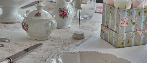 A table set with white porcelain tea service, including a teapot, sugar bowl, creamer, and a cup. A floral-patterned box is also visible.