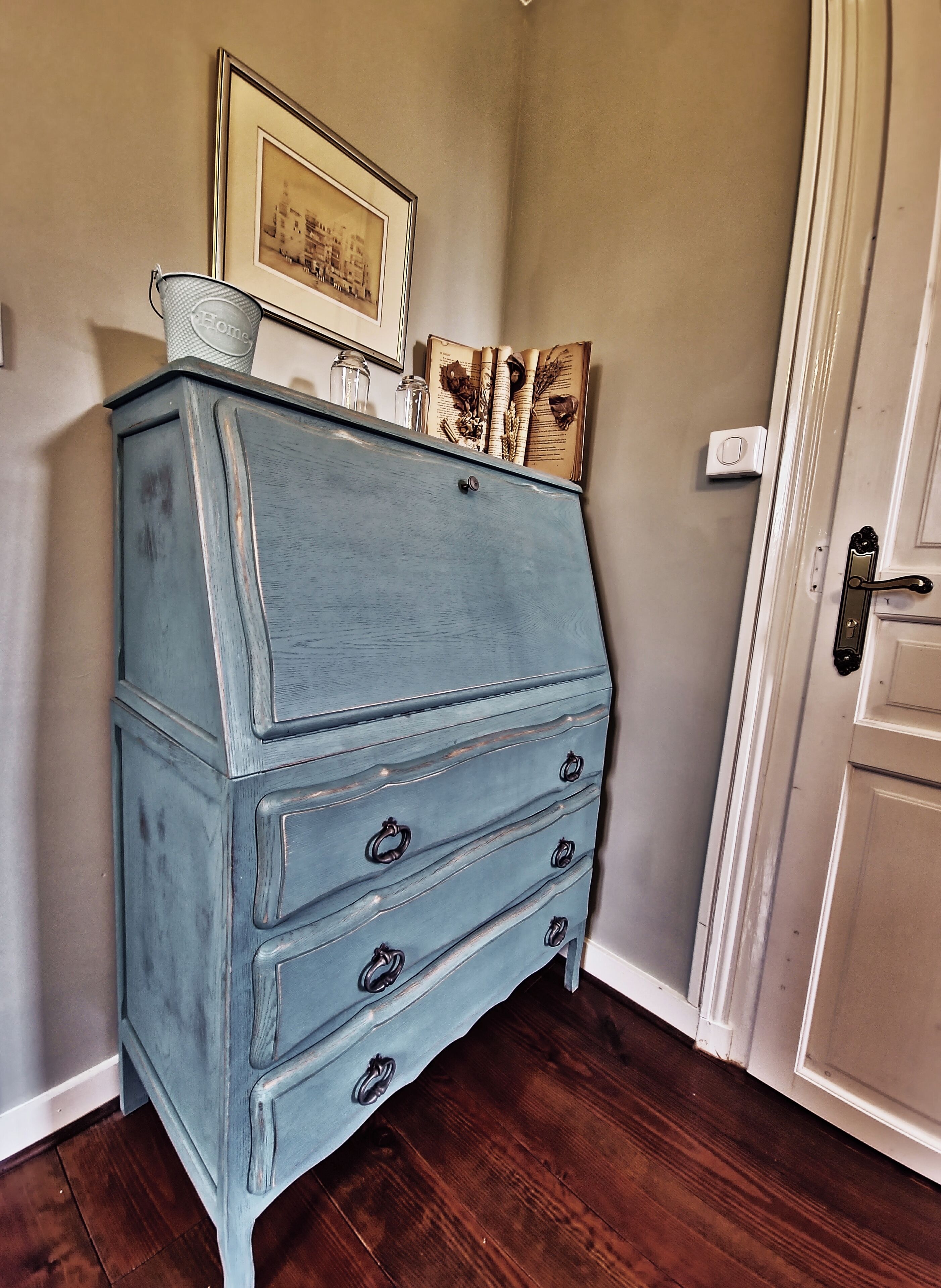 A blue dresser with three drawers, a small basket, and a framed picture on top.