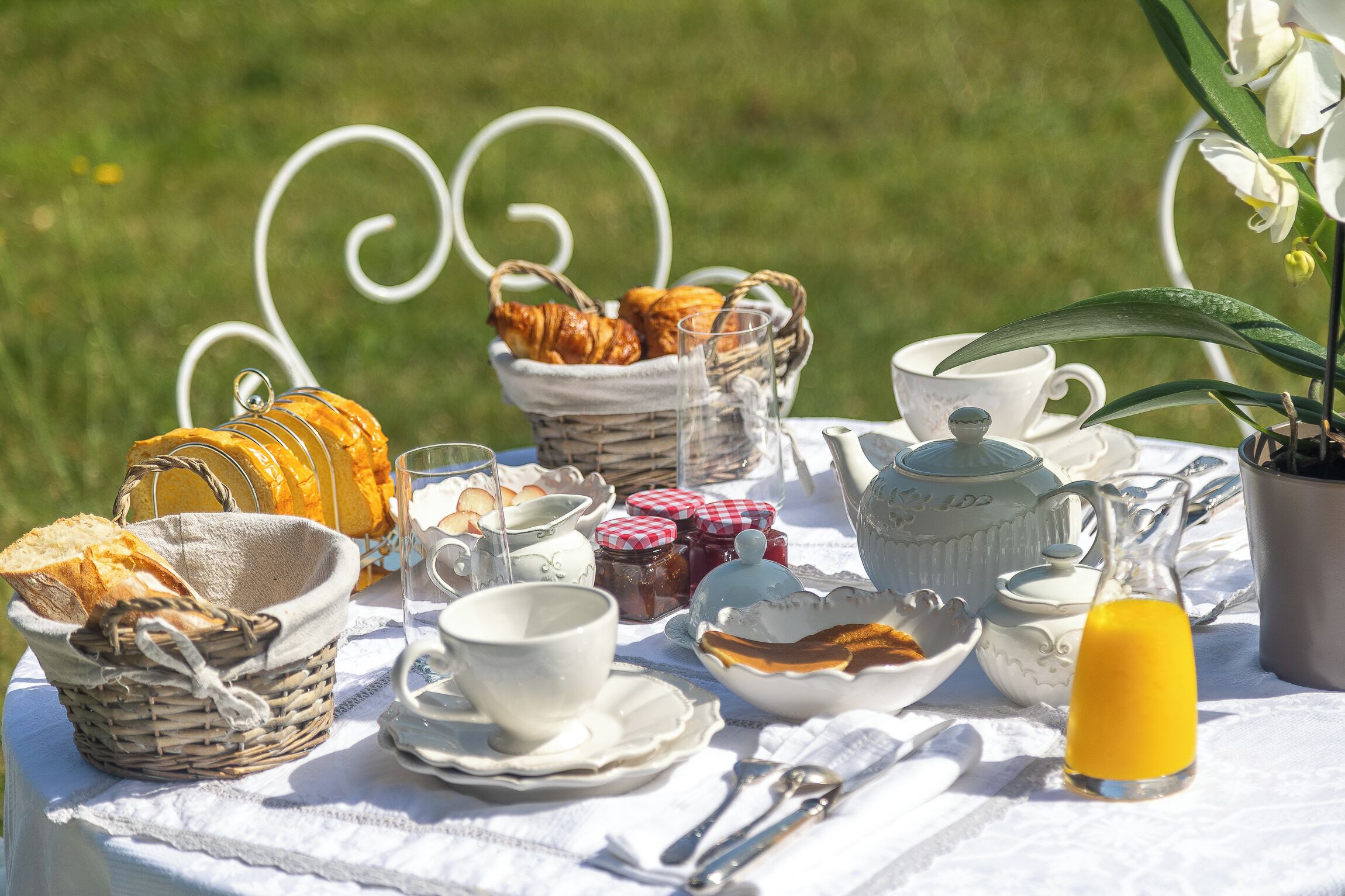 A picnic setup with bread, pastries, cups, and a jug of juice on a white tablecloth.