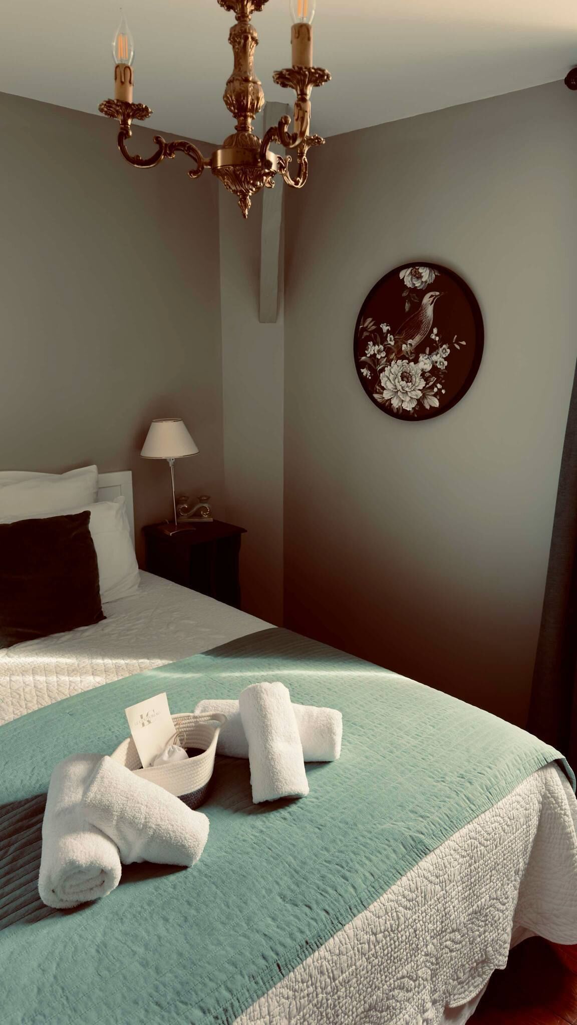 A neatly made bed with a teal and white quilt, a round wall decoration, a bedside lamp, and a chandelier.