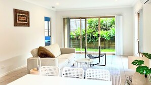 A modern living room with a sofa, chairs, and a dining table. There is a sliding glass door leading to a garden with plants.