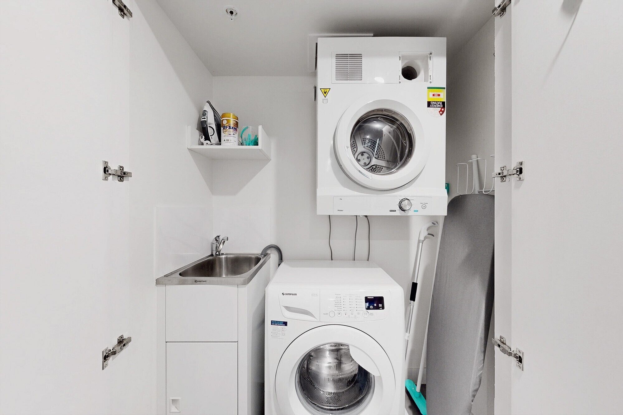 A compact laundry room with two front-loading washing machines, a sink, and a small shelf with toiletries.