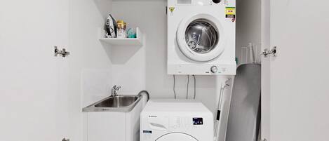 A compact laundry room with two front-loading washing machines, a sink, and a small shelf with toiletries.