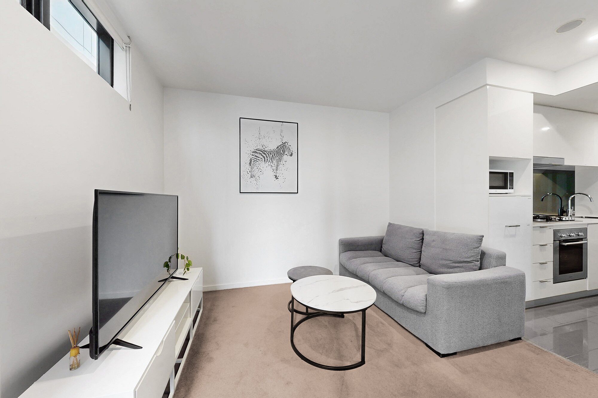 A modern living room with a grey sofa, a small round coffee table, a flat-screen TV, and a kitchen area with a microwave and oven.