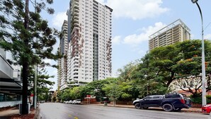 A modern high-rise building with multiple balconies, surrounded by trees and parked vehicles on a city street.