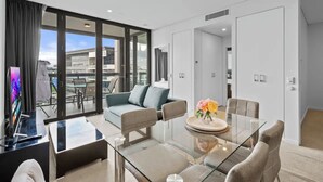 A modern hotel room with a glass dining table, a sofa, and a TV.